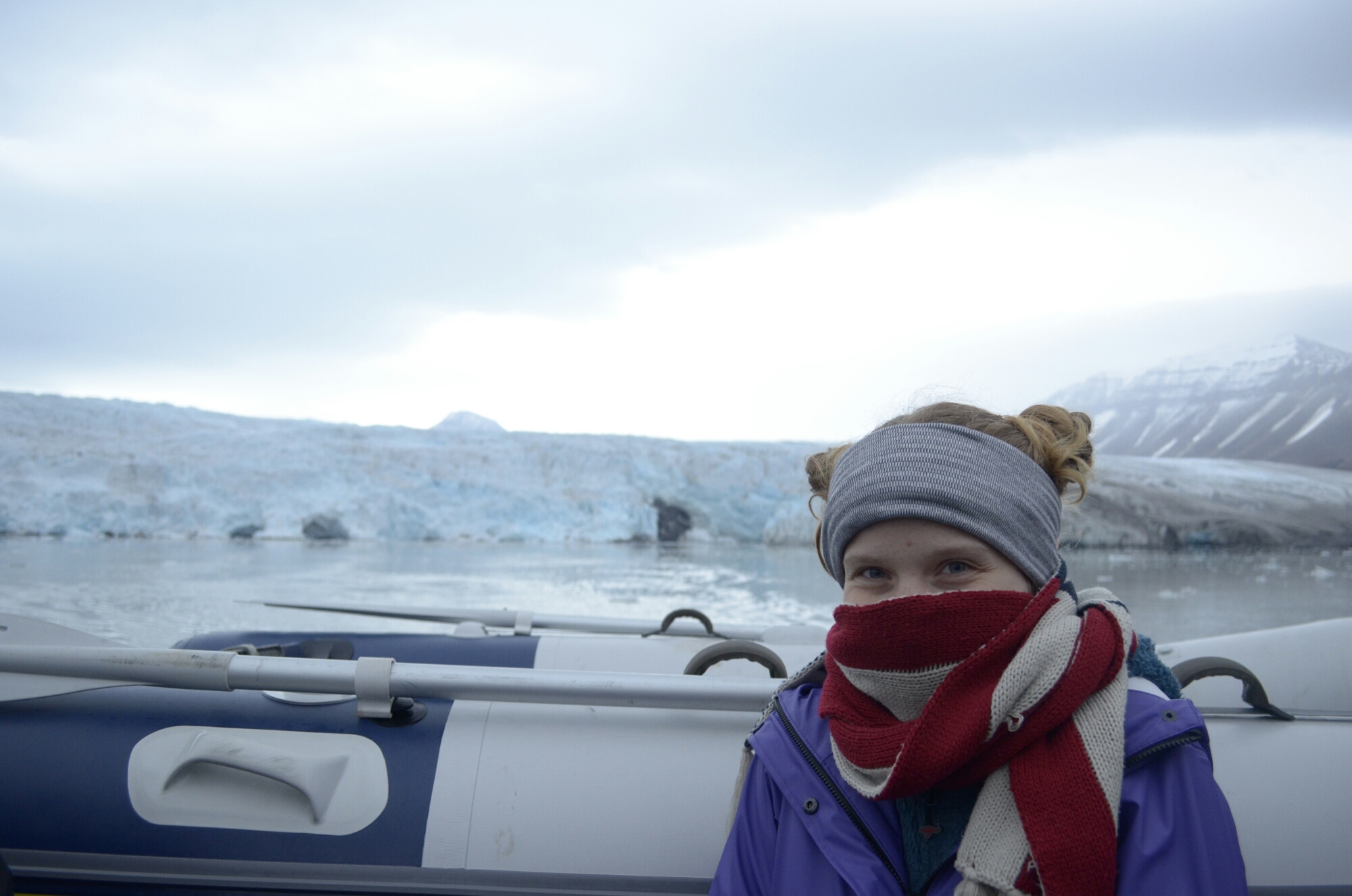a girl on a boat clearly freezing but happy in front of a glacier