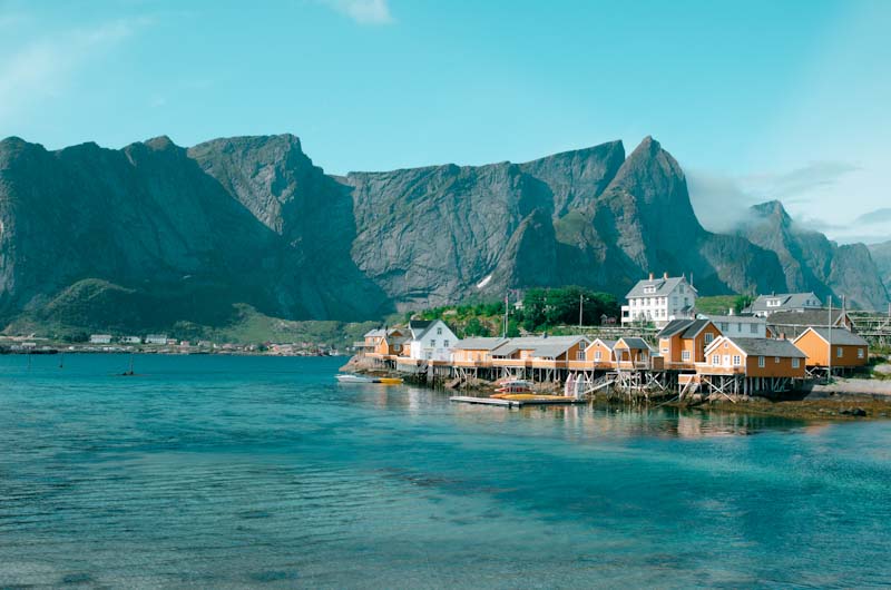 Eine Stadt aus gelben Holzhäusern auf Stelzen und einer Insel im türkisen Fjord mit Bergen im Hintergrund.